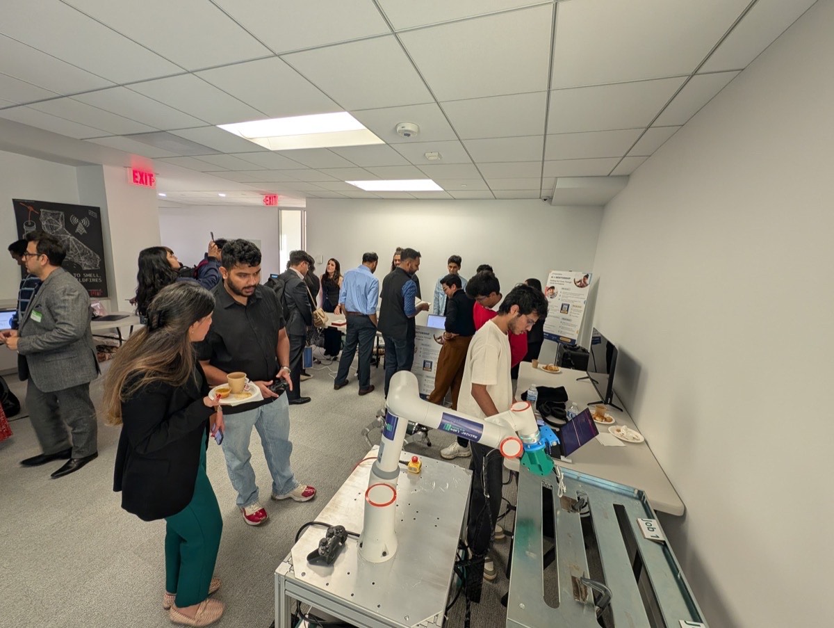The demo floor at AI Kiran: a robotic arm on a table, attendees with food and drinks, WYWA poster on the wall